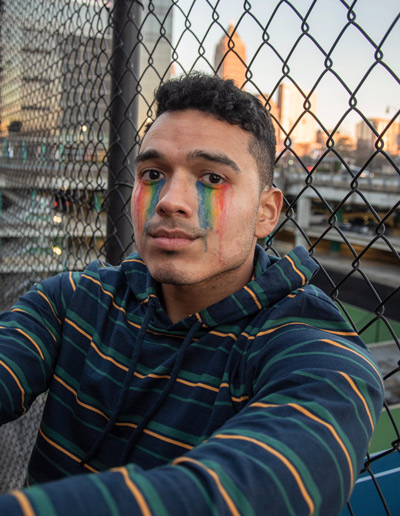 Photo: a young, Black, Latinx, LGBTQ+ person sitting against a chain-link fence with makeup of rainbow tear stains.